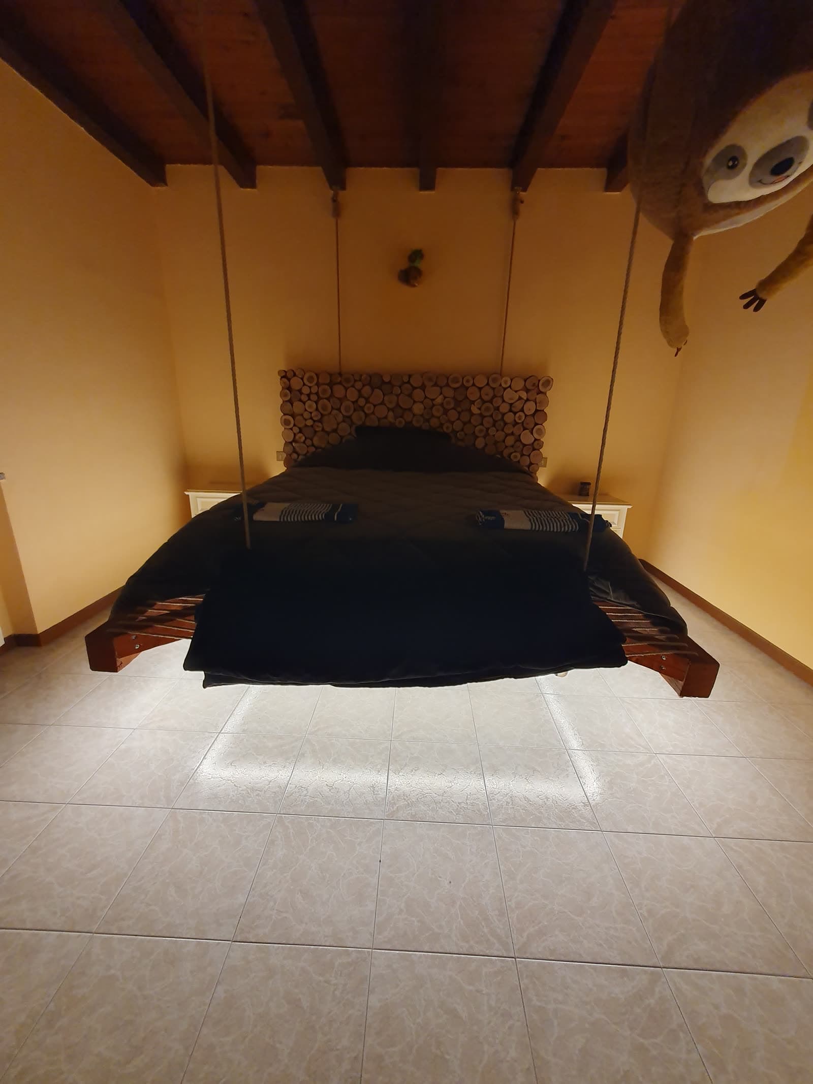Letto matrimoniale sospeso con corde dal soffitto in legno di una mansarda, con luce radente sul pavimento